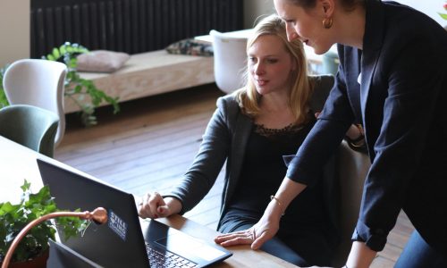 Two professional women discuss a project using a laptop in a modern office environment.